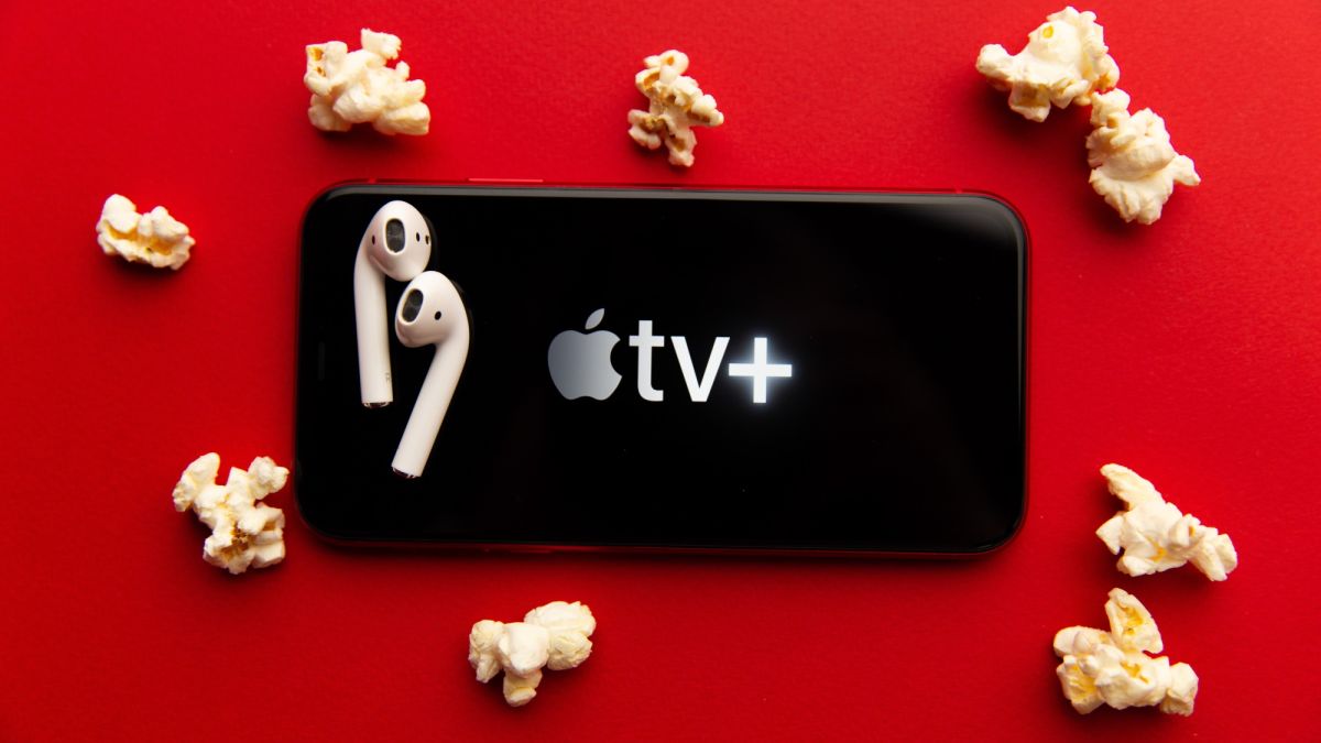 iPhone with Apple TV Plus logo, a pair of AirPods on top, laying on a red background with pieces of popcorn surrounding it