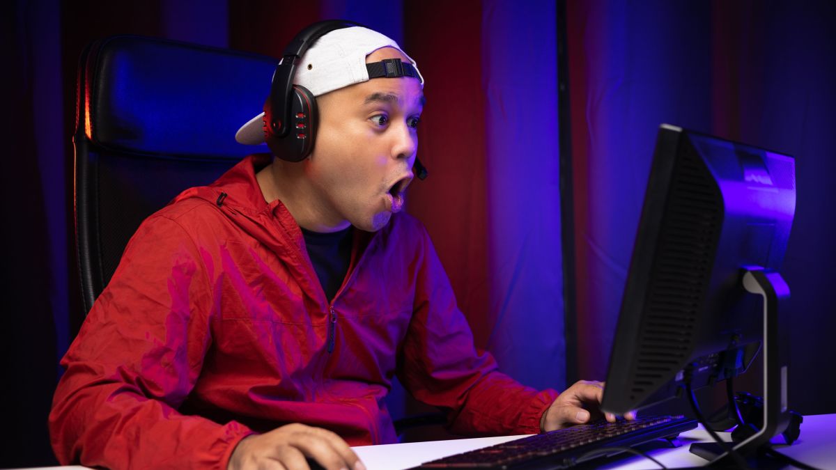 Excited and shocked face of gamer with headphone playing computer pc video game online sitting on chair at living room. 
