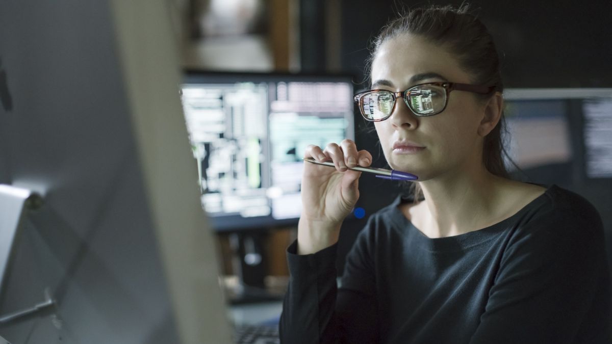 Stock photo of young woman’s face as she contemplates one of the many computer monitors that surround her.